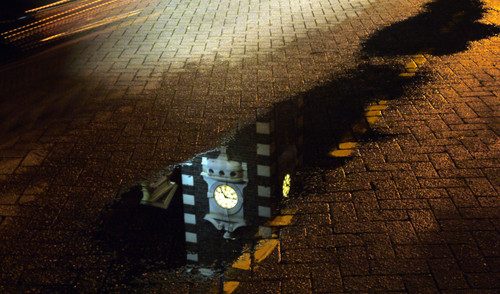 Photo is of a puddle with the reflection of the clock tower of the Railway Station at night