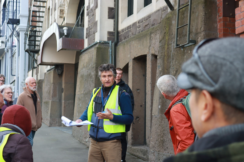 City Walks guide Athol Parks speaks in front of the fine Dowling Street facade of Garrison Hall. Visible behind him are very large blocks of Port Chalmers breccia at street and basement level. White Oamaru stone facings are visible above.