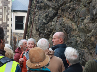 Geologist Dr Nick Mortimer stands in the Burlington Street cutting and explains the 1860s reduction of Dunedin's Bell Hill. Behind Nick is the exposed rock face, and the rear wall of Garrison Hall - an 1870s building that features this stone along with lighter-coloured Port Chalmers breccia and Oamaru stone facings. Attentive participants in the tour look on. 