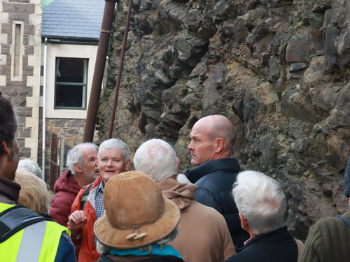 Geologist Dr Nick Mortimer stands in the Burlington Street cutting and explains the 1860s reduction of Dunedin's Bell Hill. Behind Nick is the exposed rock face, and the rear wall of Garrison Hall - an 1870s building that features this stone along with lighter-coloured Port Chalmers breccia and Oamaru stone facings. Attentive participants in the tour look on. 
