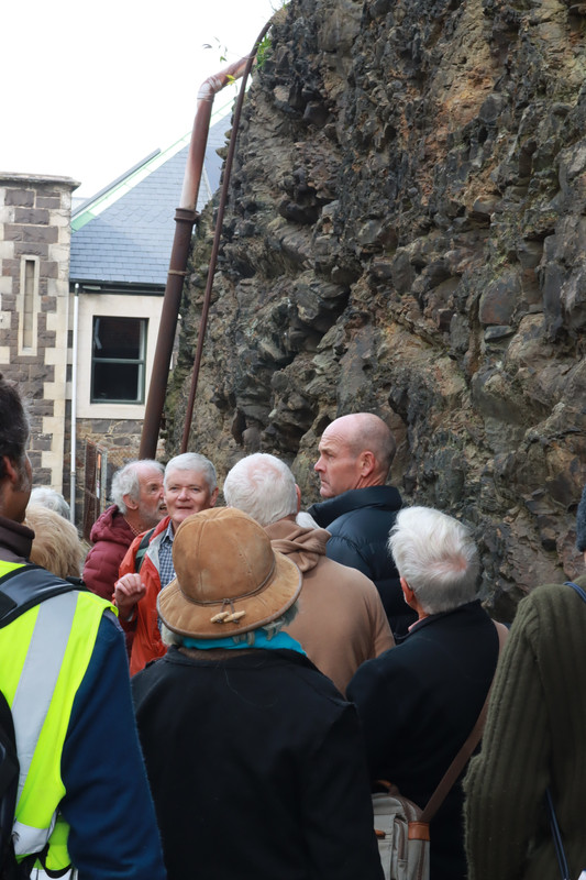 Geologist Dr Nick Mortimer stands in the Burlington Street cutting and explains the 1860s reduction of Dunedin's Bell Hill. Behind Nick is the exposed rock face, and the rear wall of Garrison Hall - an 1870s building that features this stone along with lighter-coloured Port Chalmers breccia and Oamaru stone facings. Attentive participants in the tour look on. 