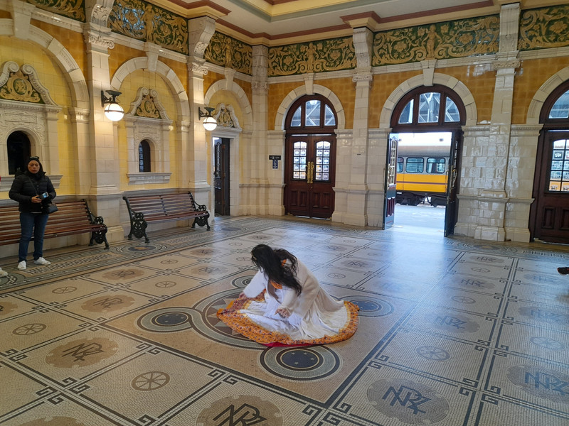 A woman in a gold-braided dress swoons on the mosaic floor of the ornate booking hall of Dunedin Railway Station.